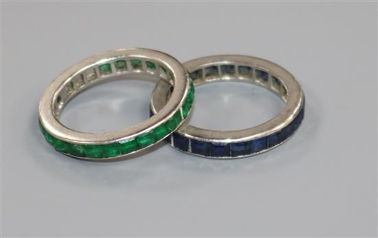 An emerald channel-set eternity ring, white metal setting (tests as platinum, two stones missing) and a similar sapphire ring